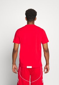 Hombre con cabello corto y rizado, vistiendo una camiseta roja sencilla y pantalones cortos deportivos rojos con ribetes blancos, de pie frente a una pared blanca.