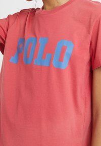 Bavlněné tričko v korálové barvě s krátkými rukávy, které nese výrazný modrý text "POLO". Hladká textura; volný střih, kulatý výstřih.