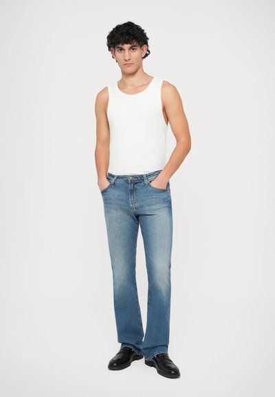 Weißes Tanktop und hellblaue Jeans aus Denim mit verblasstem Finish, stehendes Modell mit Händen in den Taschen, schwarze Schuhe, einfarbiger Hintergrund.