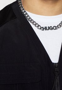 Srebrny naszyjnik z łańcuszkiem z literami "HUGO" noszony na białej koszuli i czarnej kurtce zapinanej na zamek z klapką na kieszeni.