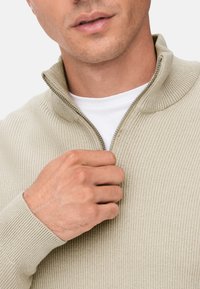 Beżowy sweter z zamkiem na szyi o ryżowej fakturze, z zabezpieczonym zamkiem i białą koszulą pod spodem. Dłoń trzymająca zamek w okolicy szyi.