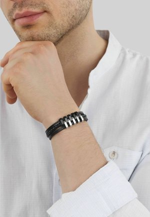 Mann in weißem Hemd, der das Kinn auf die Hand stützt und ein schwarzes Lederarmband mit silbernen Metallakzenten am Handgelenk zeigt.