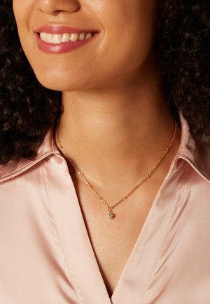 Collier en or avec une chaîne délicate et un petit pendentif rond comportant une pierre vert clair. Porté avec un chemisier en satin rose pâle.