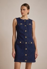 Robe sans manches en denim bleu foncé avec des fermetures à boutons dorés, deux poches avant et des coutures contrastées le long des bords.