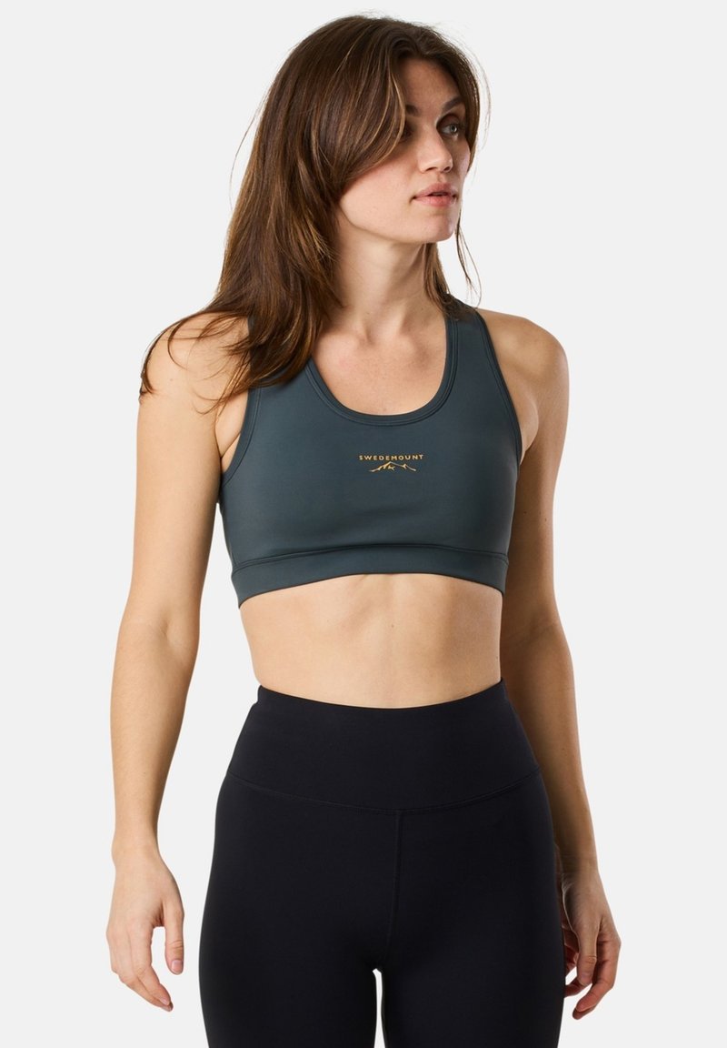 Sport-BH i mörkgrön material med rundad halsringning och guld-logo. Matchas med högmidjade svarta leggings.