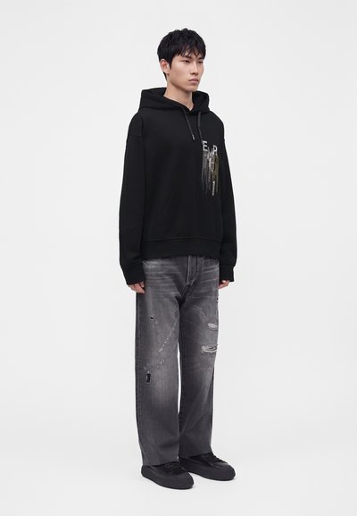 Jeune homme portant un sweat à capuche noir avec un imprimé graphique, un jean gris déchiré et ample, et des baskets noires, debout devant un fond uni.