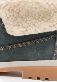 Botte en daim vert avec un revers en peau lainée crème, présentant des coutures beiges et une semelle extérieure à traction avec un motif texturé. Logo embossé sur le côté.
