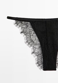 Perizoma in pizzo nero con motivo floreale trasparente, bordi ondulati e una cinta in tessuto liscio. Frange delicate lungo i lati per aggiungere un tocco di dettaglio.