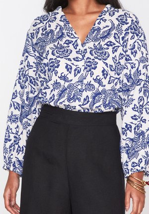 Femme portant une blouse blanche à manches longues avec un imprimé floral et d'oiseaux bleu, rentrée dans un pantalon noir taille haute, avec des bracelets dorés au poignet droit.