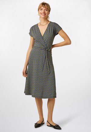 Sukienka kopertowa w niebiesko-żółty geometryczny wzór, z krótkimi rękawami, dekoltem w serek i paskiem w talii. Do tego czarne płaskie buty.