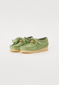 Des chaussures en daim vert avec une semelle à talon bas, des glands décoratifs, un système de laçage avec des accents en bois et des détails cousus.