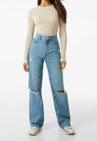 Kvinna som bär högmidjade ljusblå ripped breda byxor i jeans, kombinerat med en åtsittande beige långärmad tröja och sneakers.