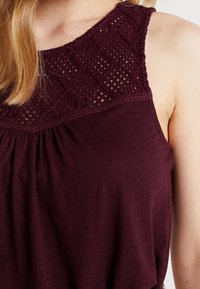 Burgunderröd ärmlös topp med en texturerad spetskrage i diamantmönster, mjukt tyg och samlad detalj vid midjan.