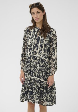 Kobieta o długich brązowych włosach, ubrana w granatowo-białą długorękawową sukienkę w kwiaty, z okularami przeciwsłonecznymi spoczywającymi na głowie, na jednolitym tle.