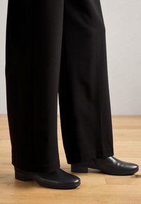 Black ankle boots with a rounded toe and low block heel, paired with wide-leg black trousers, on a wooden floor background.
