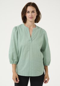 Blouse vert menthe avec un décolleté en V, manches bouffantes trois-quarts, tissu texturé présentant de petits motifs en relief et une coupe décontractée.