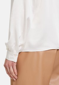 Blusa bianca in satin con maniche lunghe, polsino arricciato con bottone, abbinata a pantaloni in pelle sintetica color cammello, texture liscia, design aderente.