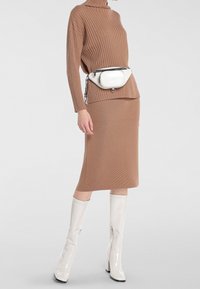 Pull en col roulé brun côtelé, jupe ajustée longueur genoux, bottes blanches brillantes jusqu'aux genoux, et un sac banane blanc avec une fermeture éclair noire.