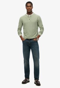 Camicetta henley verde a maniche lunghe con tre bottoni, abbinata a jeans blu in denim e scarpe marrone scuro, in piedi su uno sfondo bianco.