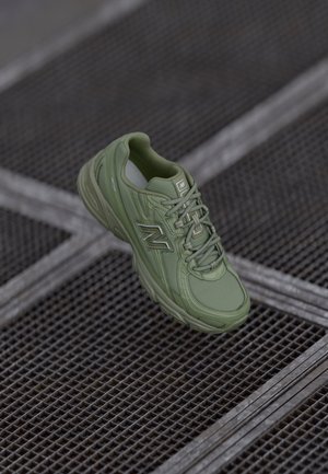 Zielony sneaker New Balance z sznurowadłami unoszący się nad ciemną metalową kratką podłogi z przecinającymi się srebrnymi liniami.