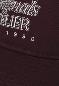 Gorra burdeos con texto bordado en blanco "Originals Atelier" y "1990". Presenta una tela suave y una visera curva, mostrando una leve textura.