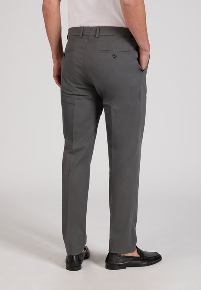 Homme portant un pantalon de costume gris ajusté et des mocassins noirs, debout, les mains dans les poches, sur un fond uni.