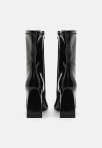 Botine din piele lăcuită neagră, cu toc gros înalt, închidere cu fermoar și un finisaj neted, lucios. Prezintă cusături decorative.