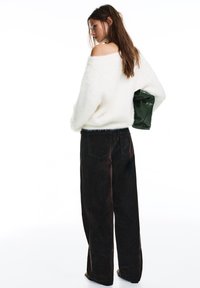 Kobieta w białym, miękkim swetrze z odkrytymi ramionami, ciemnych, szerokich spodniach, trzymająca zieloną torebkę za plecami, zwrócona bokiem.