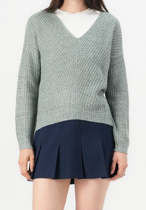 Femme portant un pull en maille gris clair à col en V par-dessus un haut blanc et une jupe plissée bleu marine, debout devant un fond uni.
