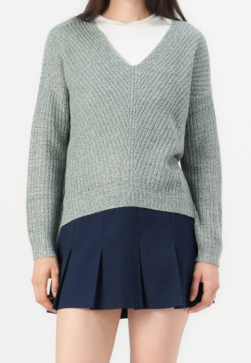 Femme portant un pull en maille gris clair à col en V par-dessus un haut blanc et une jupe plissée bleu marine, debout devant un fond uni.