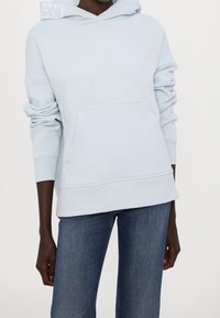 Světle modrá bavlněná mikina s kapucí, přední kapsou, žebrovanými manžetami a decentním logem na kapuci. Spárována s tmavě modrými džínami.