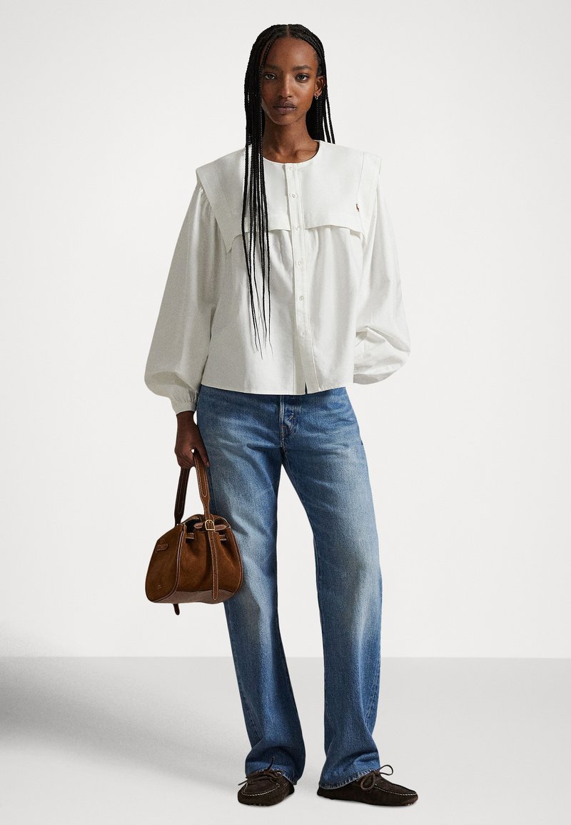 Vrouw met lange vlechten, gekleed in een witte blouse met brede kraag, blauwe spijkerbroek, donkere mocassins en een bruine suède handtas vasthoudend.