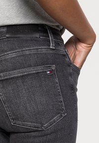 Primer plano de una persona que lleva puesta unos jeans gris oscuro de Tommy Hilfiger, con la mano en el bolsillo delantero y el logotipo de la marca visible en la cintura trasera y el bolsillo.