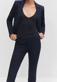 Personne portant un blazer bleu marine et un pantalon avec un haut noir en V, tenant la taille du pantalon avec les deux mains.