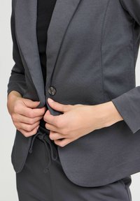 Szara marynarka o gładkiej fakturze, zapinana na jeden guzik, z klapowym kołnierzem. Szczegóły obejmują subtelny połysk i dopasowany krój.