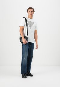 T-shirt blanche avec un logo graphique gris, jean en denim bleu avec une coupe décontractée, sac crossbody noir et chaussures en cuir noires.