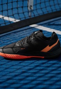 Μαύρο αθλητικό παπούτσι με υφή στην επάνω επιφάνεια, πορτοκαλί λεπτομέρειες και σόλα με σχέδιο σε ζιγκ ζαγκ. Τοποθετημένο σε μπλε επιφάνεια γηπέδου κοντά σε δίχτυ.