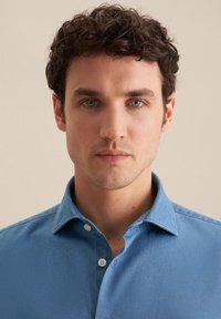 Giovane con capelli ricci castani, occhi azzurri, indossando una camicia blu con bottoni, che guarda dritto davanti a sé su uno sfondo semplice.