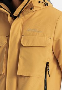 Chaqueta impermeable amarilla con un acabado texturizado, que presenta un bolsillo con cremallera y dos bolsillos de solapa en el pecho. Detalle del logotipo visible en la tela.