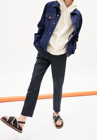 Persona vestida con una chaqueta de mezclilla azul oscura sobre una sudadera crema, pantalones negros y sandalias negras con ribete beige, de pie con una pierna hacia adelante.