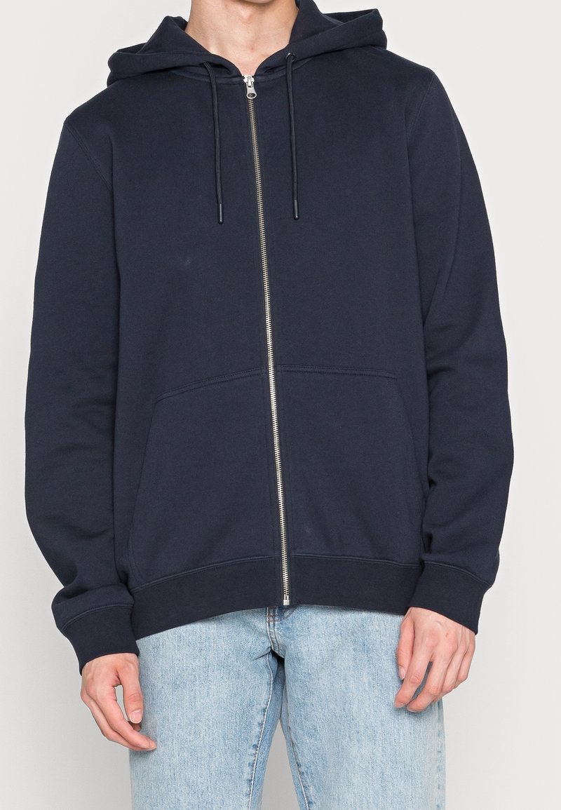 Felpa con zip navy realizzata in tessuto morbido. Presenta un cappuccio con cordino, tasche a marsupio e polsini a coste con una cerniera liscia.