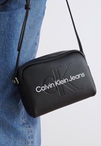 Borsa a tracolla rettangolare nera realizzata in pelle liscia, con logo in rilievo "Calvin Klein Jeans" e chiusura con zip.