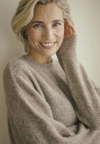 Lachende blonde vrouw met blauwe ogen, gekleed in een gebreide beige trui, die haar haar aanraakt, tegen een effen neutrale achtergrond.