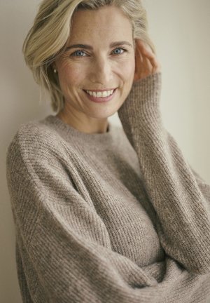 Naeratav blond naine siniste silmadega, seljas heledast beezist kudum, puudutab oma juukseid, neutraalse tausta taustal.