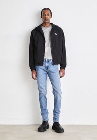 Giacca nera con cappuccio e polsini elasticizzati sopra una maglia a righe, abbinata a jeans azzurri chiari e anfibi neri voluminosi, in piedi davanti a uno sfondo neutro.