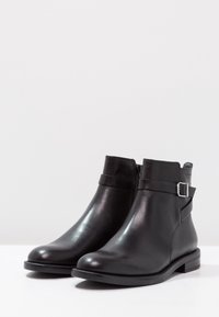 Botas de cuero negro hasta el tobillo con un acabado suave, cierre de cremallera lateral y un detalle de tira con hebilla. Diseño robusto, con tacón bajo y punta redondeada.