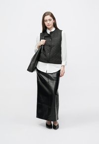 Črni prešiti telovnik preko bele srajce, kombiniran z dolgo črno usnjeno krilo in črnimi čevlji. Model drži črno torbico tote.