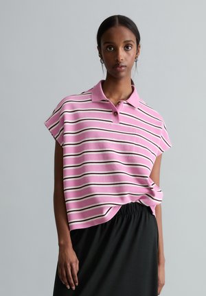 Woman wearing a pink and white striped short-sleeve polo shirt tucked into a black skirt, standing against a plain gray background.