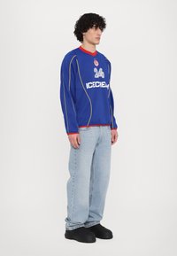 Blauw longsleeve shirt met gele accenten en rode kraag, met het 'ICECREAM' logo en het nummer '24' op de voorkant; gecombineerd met lichtblauwe jeans en zwarte schoenen.