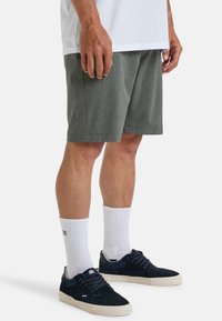 Shorts verts en tissu texturé, avec une coupe décontractée et des poches latérales, associés à des chaussettes blanches et des baskets bleu marine.